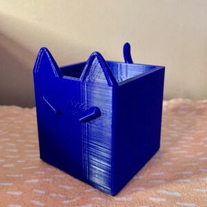 Puede incluir: Un macetero de gato azul impreso en 3D. El macetero tiene una base cuadrada y una cabeza de gato en la parte delantera con orejas y cola.