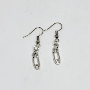 Puede incluir: Un par de pendientes plateados. Cada pendiente presenta un pequeño dije de imperdible suspendido de un anzuelo. Los dijes de imperdible tienen una cabeza redondeada y un cierre.