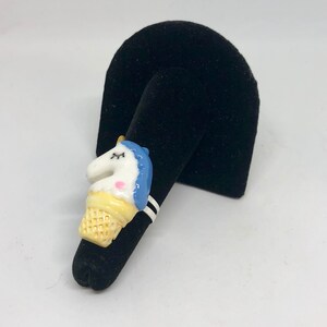 Könnte beinhalten: Ein Ring in Form eines Eisbechers mit einem weißen Einhorn, blauen und gelben Akzenten. Der Ring ist aus Keramik und hat ein weißes Band mit schwarzen Streifen.