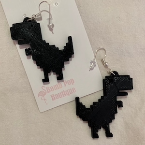 May include: A pair of black pixelated dinosaur earrings with silver hooks. The earrings are on a white card with the text "Bomb Pop Boutique" printed on it.