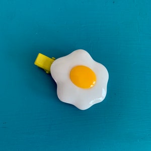 May include: A white and yellow fried egg hair clip with a yellow clip base. The egg has a white yolk and a bright yellow yolk. The clip is set against a teal blue background.