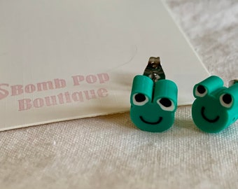 May include: A pair of teal green frog stud earrings with white eyes and a black smile. The earrings have silver posts.