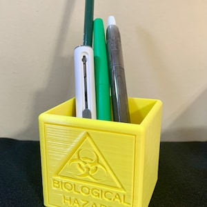 May include: Yellow desk organiser with a biohazard symbol and the text "BIOLOGICAL HAZARD". The organiser is filled with a green pen, a white pen, and a pencil.