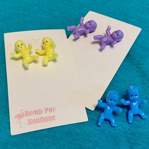 Può includere: Tre paia di orecchini a forma di bambola. Un paio è giallo, un paio è viola e un paio è blu.