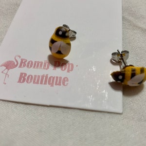 Puede incluir: Un par de pendientes de botón plateados con un dije de abeja amarillo y negro. El dije de abeja tiene rayas negras y alas blancas.