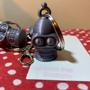 May include: A gray 3D printed keychain featuring a smiling robot head with large eyes. The keychain is attached to a silver key ring. The keychain is on a red and white polka dot background with a white card that says "Bomb Pop Boutique".