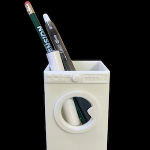 以下が含まれることがあります： 洗濯機型の白いペン立て。丸いドアがあり、上には「Lavello」の文字。中には緑の鉛筆、白いマーカー、黒いペンが入っています。