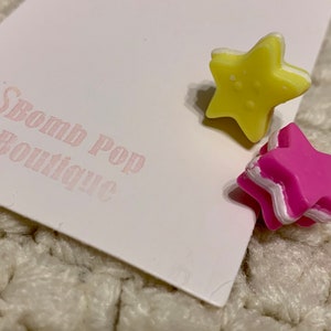 Könnte beinhalten: Zwei sternförmige Ohrringe aus Polymer Clay. Ein Ohrring ist gelb mit weißem Rand und der andere ist pink mit weißem Rand.