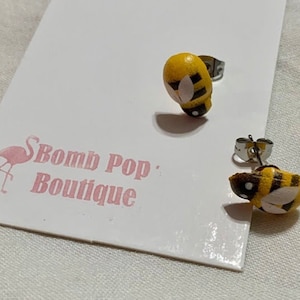 May include: A pair of silver stud earrings with a yellow and black bee design. The bee is painted with white wings and black stripes. The earrings are on a white card with the text "Bomb Pop: Boutique" and a pink flamingo.