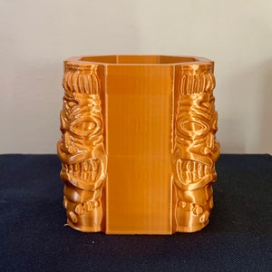 Pode incluir: Um vaso hexagonal de cor bronze com um design tiki. O vaso apresenta rostos tiki esculpidos nas laterais, com olhos, bocas e outras características faciais detalhadas. O vaso é feito de um material sólido e é adequado para uso interno ou externo.