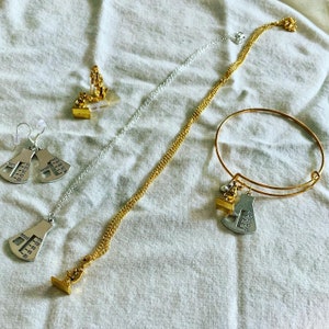 May include: Two gold necklaces with silver charms shaped like protractors, one with a silver charm shaped like a small person, and a gold bangle bracelet with a silver protractor charm. The necklaces and bracelet are on a white fabric background.