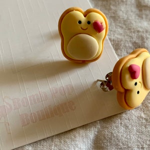 Op de afbeelding: Een paar schattige, gele en bruine toastvormige oorbellen met een roze hartje en een smiley. De oorbellen zijn op een witte achtergrond met de tekst "Strawberry Boutique".
