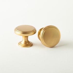 May include: Two gold-colored cabinet knobs with a round top and a pedestal base. The knobs have a brushed finish and are set against a white background. The image includes a "2 PACK" label.