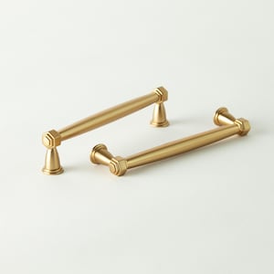 May include: Two gold-colored cabinet pulls with a cylindrical bar and hexagonal accents. The pulls are made of metal and have a matte finish. The hardware is designed for use on drawers or cabinets.