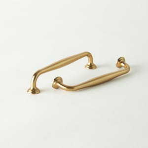 May include: Two gold-colored cabinet pulls with a curved design. The pulls have a smooth, polished finish and are set against a white background. The hardware is designed for easy installation and adds a touch of elegance to any cabinet or drawer.