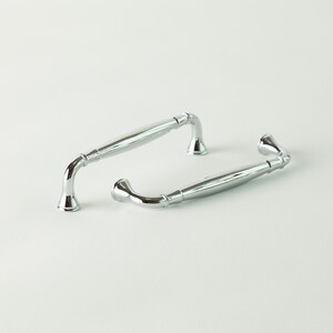May include: Two polished chrome cabinet pulls. The pulls have a curved design with a central bar and flared ends. The hardware is designed for use on drawers or cabinet doors. The shiny metal finish reflects light.