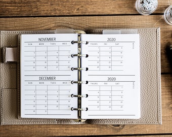 Op de afbeelding: Open planner met kalenders voor november en december 2020. De planner is lichtgrijs, met een ringband. De roosters van de maanden zijn zichtbaar. Een pen en andere kantoorbenodigdheden liggen op het houten oppervlak.