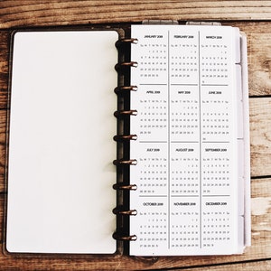 May include: Open planner with a 2019 calendar displayed. The pages are white with black text, showing months from January to December. The planner has a brown cover and a spiral binding.