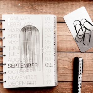 May include: A white planner with a black spiral binding. The planner has a monthly calendar view with the months January through December listed down the left side. The numbers 01 through 12 are listed down the right side. The planner is open to the month of September. The planner is on a wooden surface with a silver metal card holder and two black and white pens.