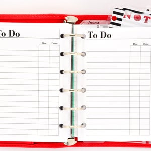 May include: A red leather bound planner with a white page open to a to-do list. The planner has a ring binder and a clear plastic pocket on the right side that holds a notepad with the word "NOTES" printed on it.