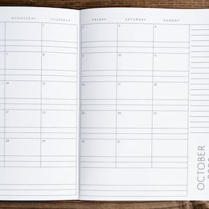 May include: A blank monthly calendar planner for October 2020. The calendar has a grid layout with spaces for each day of the month. The calendar is open to the month of October and is lying flat on a wooden surface.
