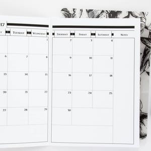 May include: A black and white monthly calendar planner for November 2017. The calendar has a grid layout with each day of the month labeled. The calendar is open to the month of November and is lying flat on a white surface.