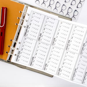 May include: A white calendar with a black spiral binding. The calendar is open to the year 2018 and shows the months of January through December. A red pen is lying on top of a brown folder.