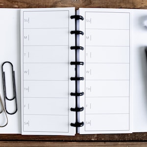 May include: A white weekly planner with black lines and the days of the week labeled: Sunday, Monday, Tuesday, Wednesday, Thursday, Friday, Saturday. The planner is open to a blank week. A black pen and two paper clips are on the left side of the planner.