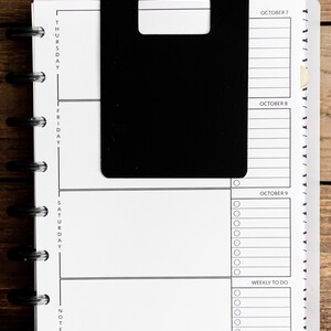May include: A black and white planner with a black tab on the left side. The planner has sections for Thursday, Friday, Saturday, and notes. The dates October 7, 8, and 9 are listed on the right side of the planner. The planner has a section for weekly to do items.