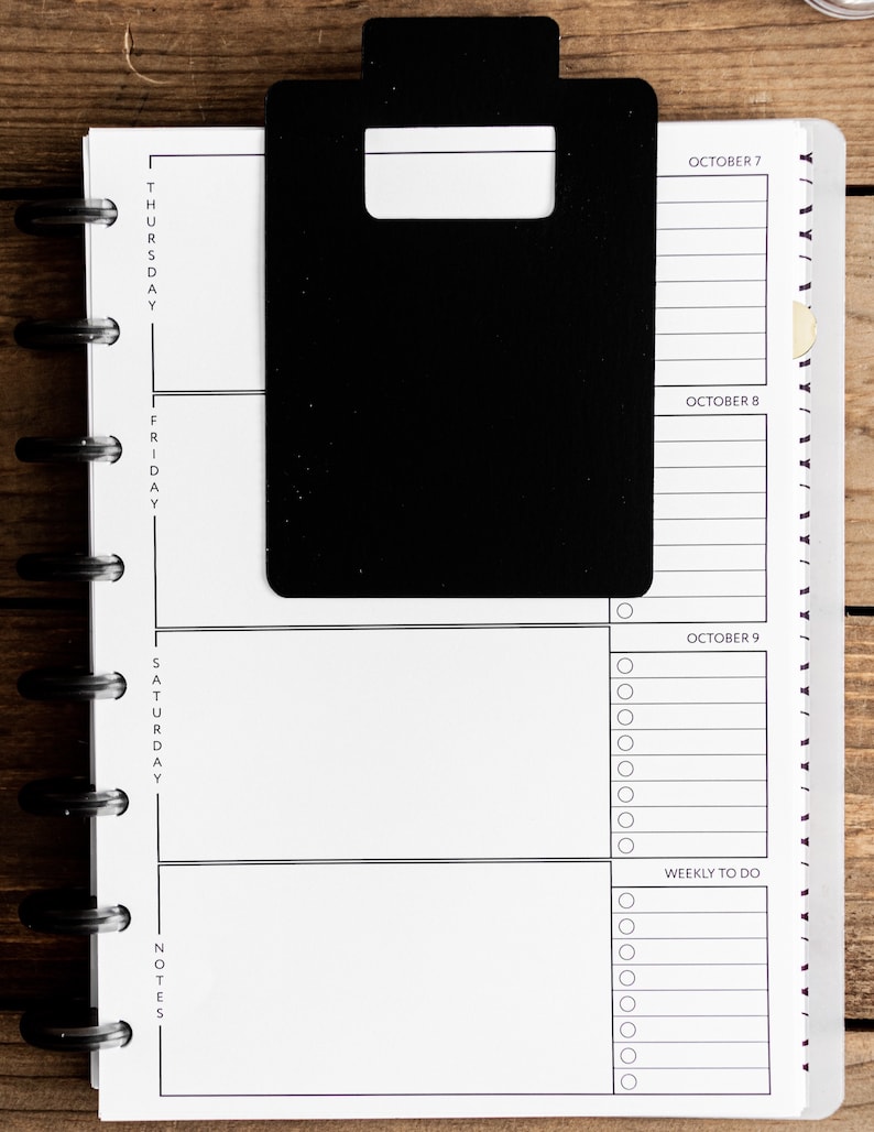 May include: A black and white planner with a black tab on the left side. The planner has sections for Thursday, Friday, Saturday, and notes. The dates October 7, 8, and 9 are listed on the right side of the planner. The planner has a section for weekly to do items.