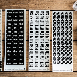 May include: Four sheets of black and white stickers with different designs for a planner or journal. The stickers include numbers, days of the week, and abbreviations for the days of the week. The stickers are on a wooden surface.