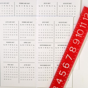 May include: A red ruler with white numbers from 3 to 12 is placed diagonally over a set of 12 monthly calendars for the year 2017.