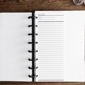 May include: An open notebook with a black binder and white pages. The right page has the word "NOTES" at the top and lined paper below. A black pen is lying on a wooden surface next to the notebook.