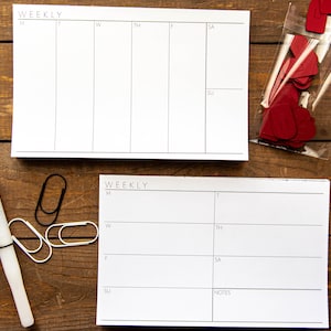 May include: Two white weekly planner pads with black lines and text. The pads are labeled "WEEKLY" and have spaces for each day of the week. A pen, paper clips, and red heart-shaped cutouts are also visible.