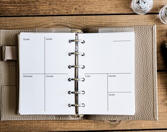 Op de afbeelding: Open weekplanner met een wekelijkse indeling, met secties voor elke dag van de week. De planner is gebonden in een leren, lichtbruine kaft. Een pen, een marker en paperclips liggen op een houten oppervlak.