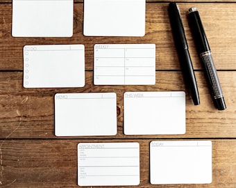 May include: A set of eight blank white note cards with different headings, including "Notes", "This Month", "To Do", "Weekly", "Memo", "This Week", "Appointment", and "Today". The cards are on a wooden surface with two pens.