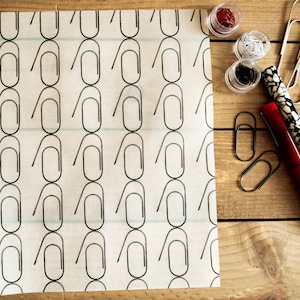 May include: White paper with a repeating pattern of black paperclip outlines. The paper is on a wooden surface with various paperclips and a red and black pen.
