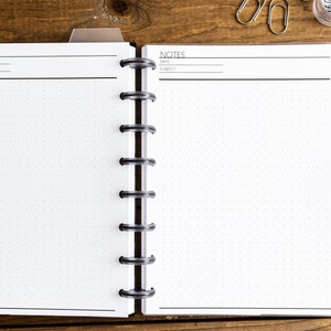 May include: An open notebook with a black and white dotted grid pattern. The notebook has a black spine with silver rings. The notebook is open to two blank pages with the word "NOTES" at the top of each page. The notebook is on a wooden surface.
