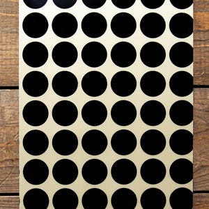May include: A rectangular metal sign with a cream-colored background and a repeating pattern of large, evenly spaced black circles. The sign has a glossy finish and is displayed against a wooden surface.