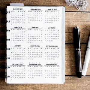 May include: A close-up of a white planner with a black spiral binding. The planner has monthly calendar pages for the year 2021. Each month is labeled with the month and year. The calendar pages are printed on white paper with black ink.