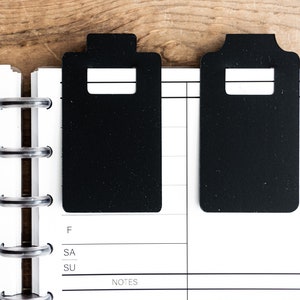 May include: Two black rectangular bookmarks with rounded corners and a rectangular cut-out in the center. The bookmarks are placed on a white page with black lines and the words "F", "SA", "SU", and "NOTES".