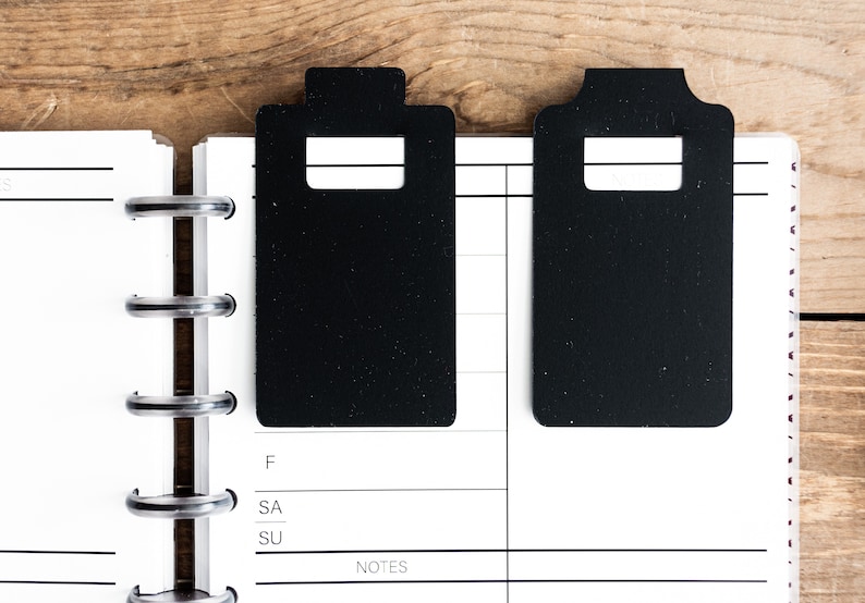 May include: Two black rectangular bookmarks with rounded corners and a rectangular cut-out in the center. The bookmarks are placed on a white page with black lines and the words "F", "SA", "SU", and "NOTES".