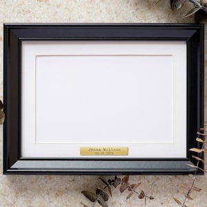 May include: A black framed picture with a white mat and a small gold nameplate. The nameplate reads "Jenna Willson 05.19.2026". The frame is rectangular and the background is a light beige.