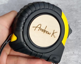 Peut inclure: Un ruban à mesurer noir et jaune avec un cercle en bois sur le devant qui dit "Andrew K."