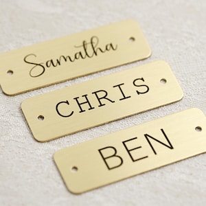 Könnte beinhalten: Drei rechteckige Namensschilder aus Messing mit den eingravierten Namen: "Samantha" in Schreibschrift, "CHRIS" in Blockbuchstaben und "BEN" in Blockbuchstaben. Jedes Schild hat zwei kleine Löcher.