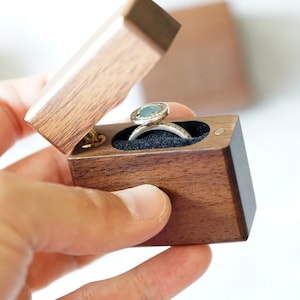 Peut inclure: Une bo&icirc;te &agrave; bague en bois avec un couvercle &agrave; charni&egrave;re, ouverte pour r&eacute;v&eacute;ler une bague avec une pierre pr&eacute;cieuse bleu clair et une bande argent&eacute;e.