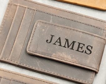 May include: A close-up of several leather cardholders in various shades of brown and gray. The central cardholder is personalized with the name "JAMES" engraved on a rectangular flap. The cardholders have multiple card slots and a distressed finish.