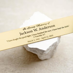 May include: A gold-colored rectangular memorial plaque with rounded edges, inscribed with the text "In Loving Memory of Jackson W. Anderson." The plaque includes a quote from 2 Timothy 4:7 and dates March 5, 1941 - April 12, 2025.