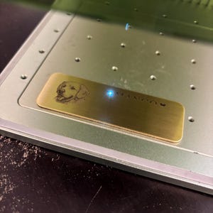 May include: A brass nameplate with a laser-etched image of a dog's face and the name "MANGO" is positioned inside a metal machine. The nameplate is rectangular and has a polished finish. The machine has a light green color.