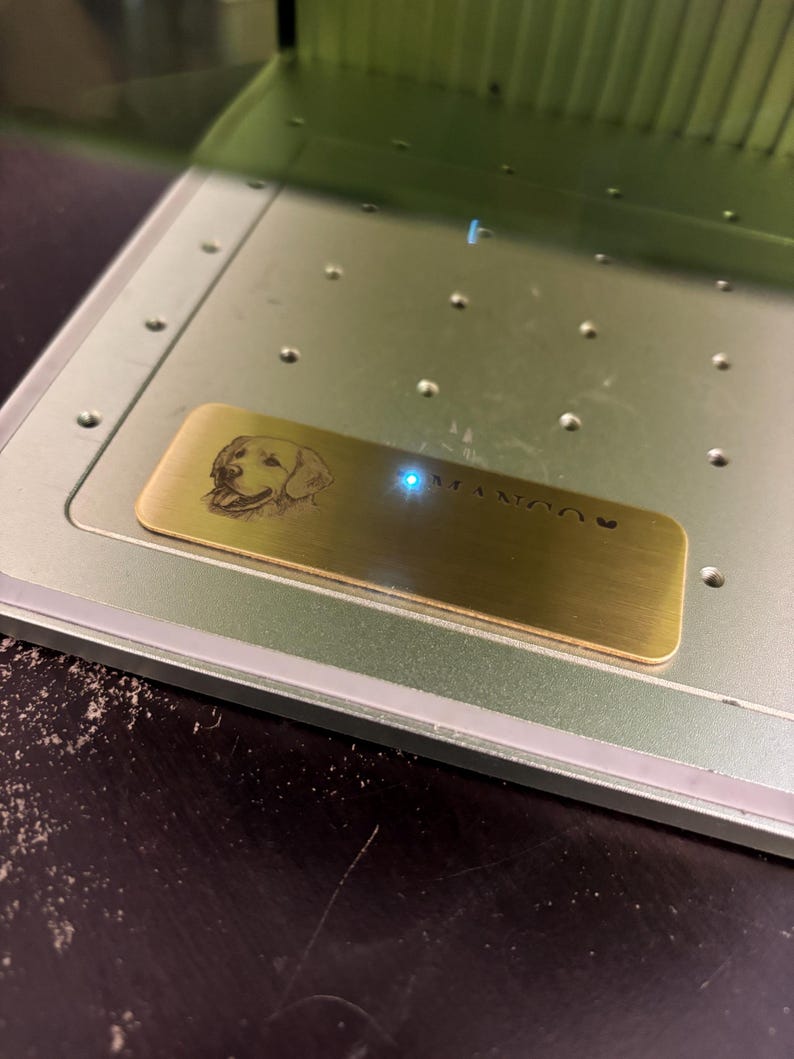 May include: A brass nameplate with a laser-etched image of a dog's face and the name "MANGO" is positioned inside a metal machine. The nameplate is rectangular and has a polished finish. The machine has a light green color.